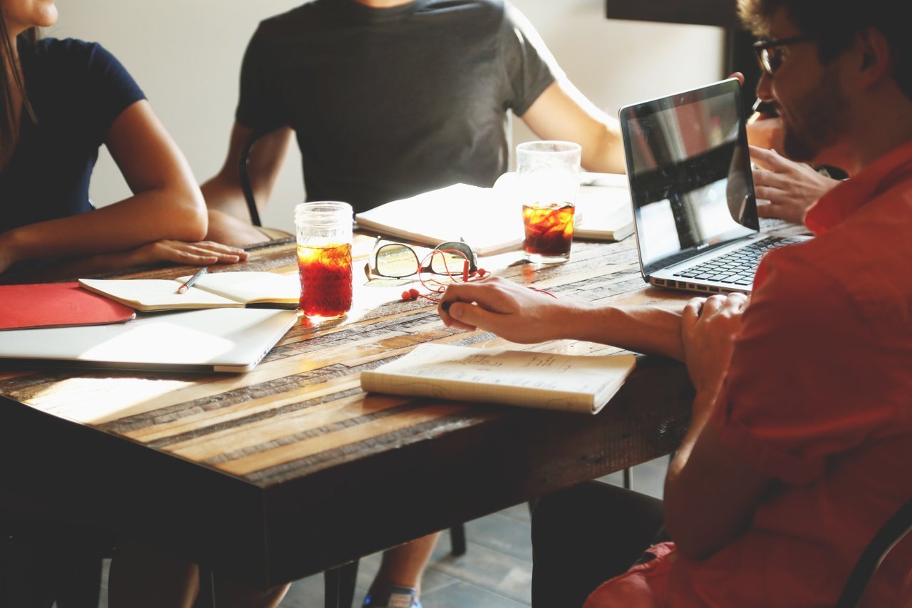 An office table with several people sitting around it and taking notes, drinking iced coffees, or typing on a laptop