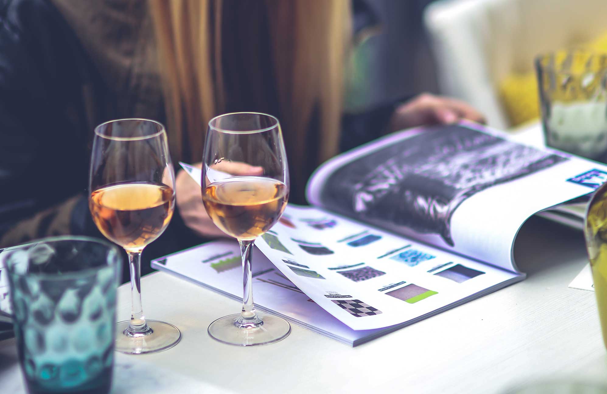 Two glasses of wine next to someone reading a magazine