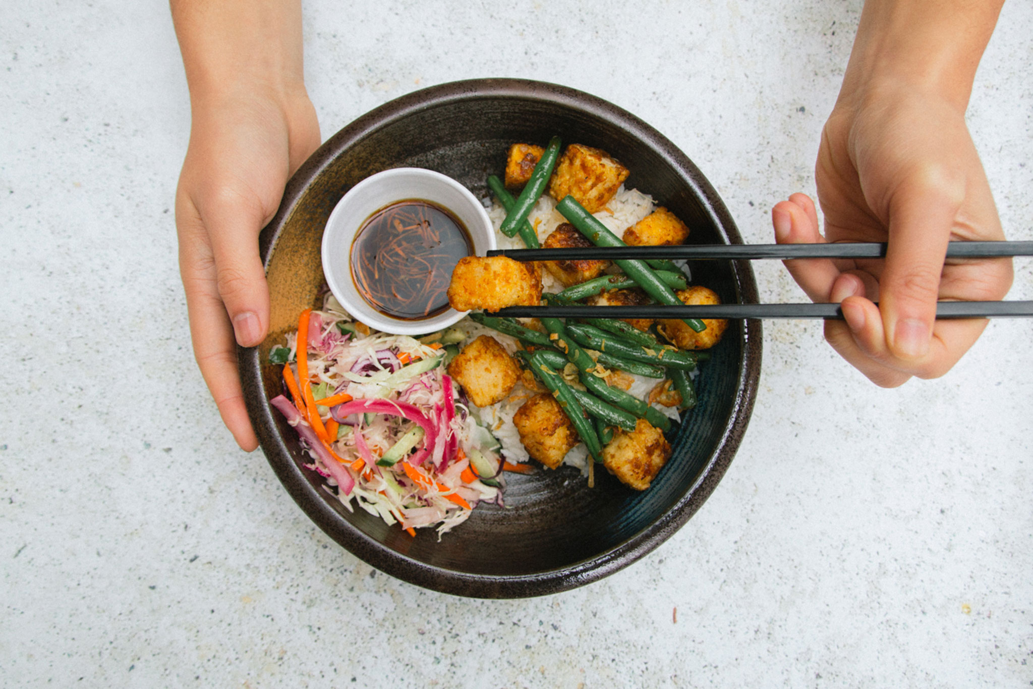 Hands holding chopsticks dig into an Asian bowl with sauce