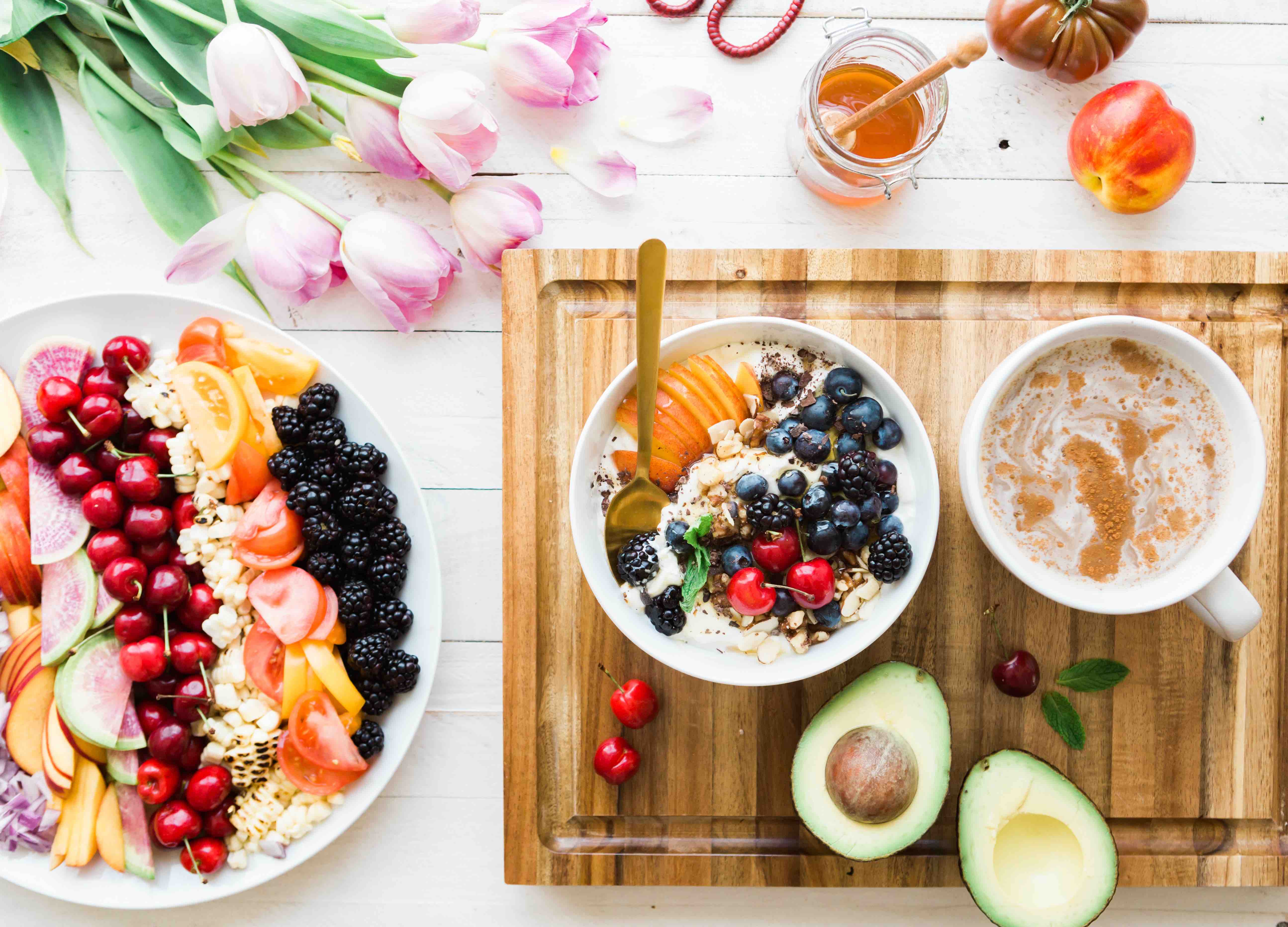Bowl of sliced fruit, flowers, a latte, granola, and avocado on a board