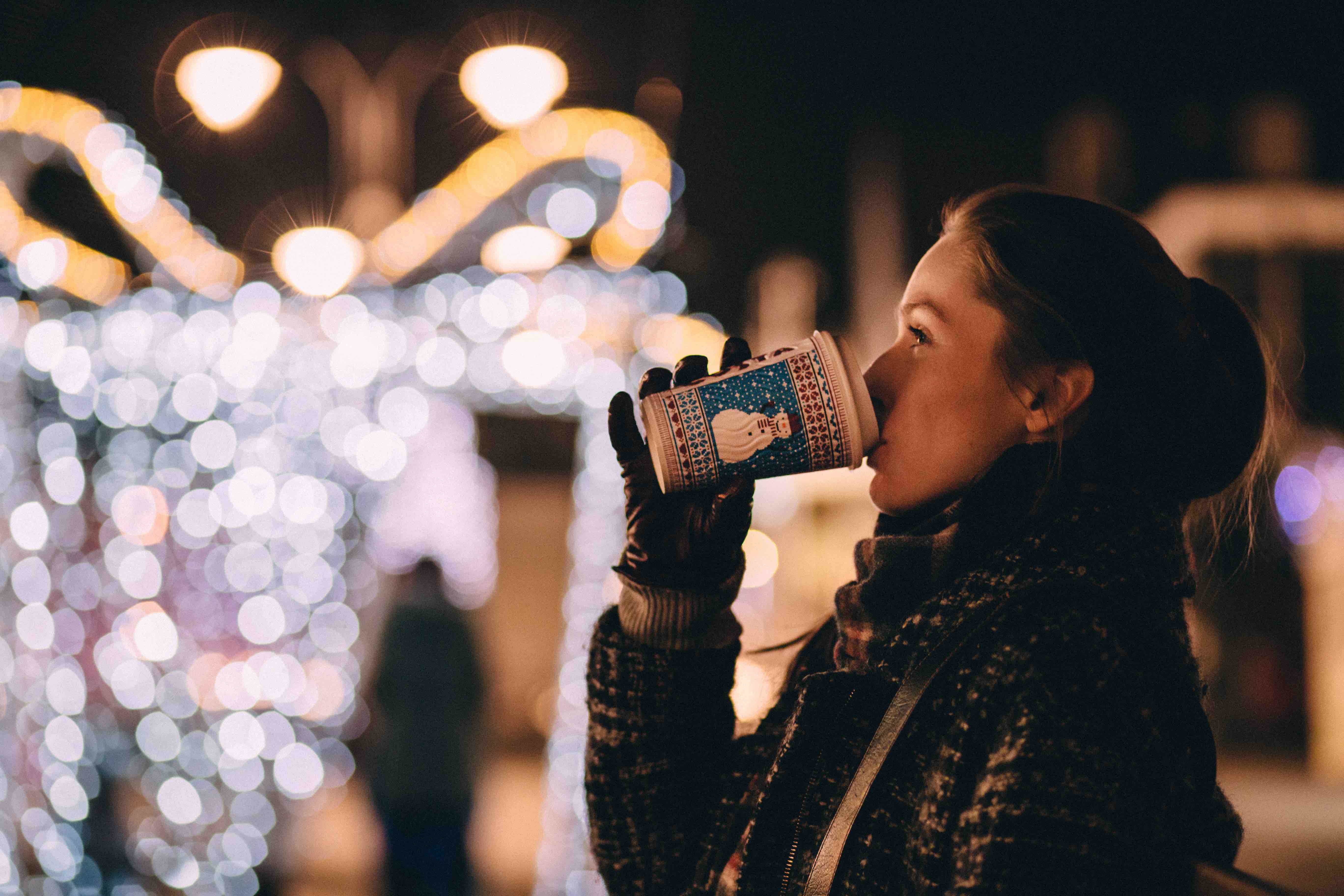 A woman sipping a holiday coffee in front of festive lights at night
