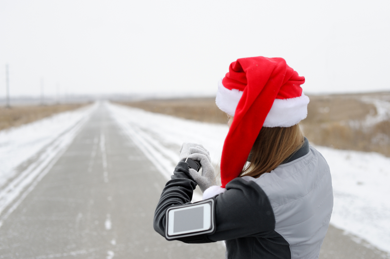 A jogger in a Santa hat on a snowy road sets their smart watch and phone