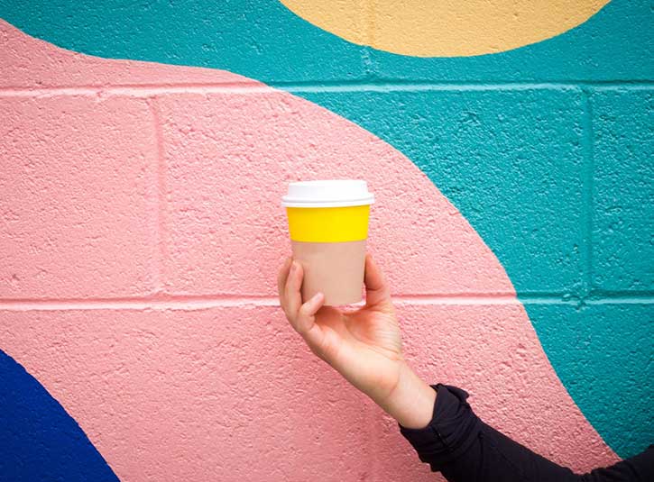 A hand holds a togo coffee in front of a painted wall