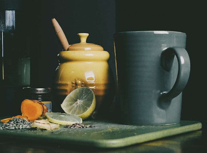 A mug of tea, lemon wedge, honey pot, and turmeric on a cutting board