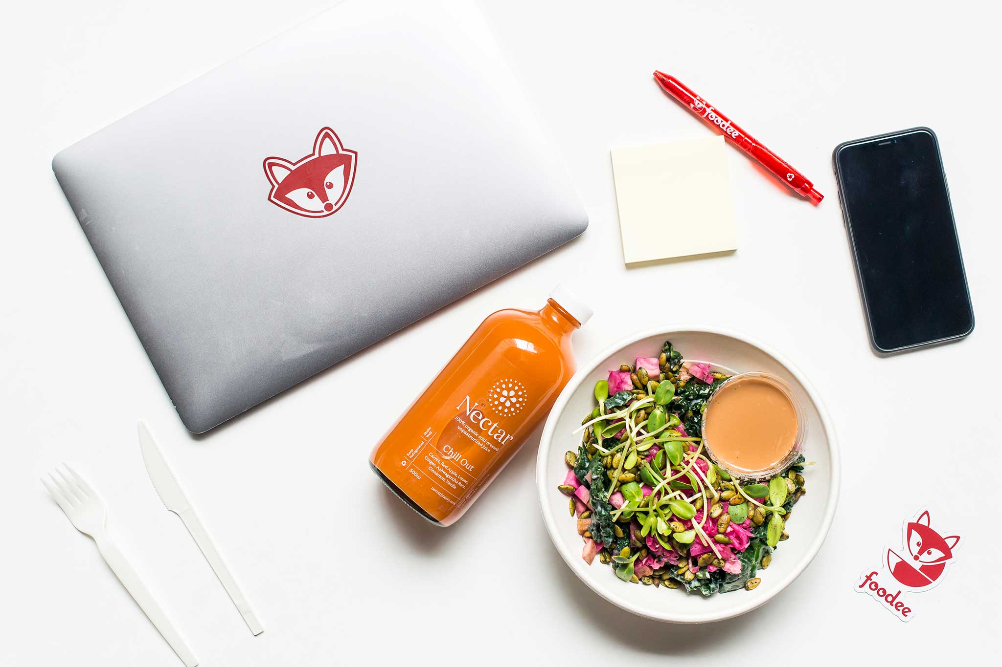 Foodee branded laptop, pen, and sticker, next to a bowl of salad, and orange juice, and a cellphone