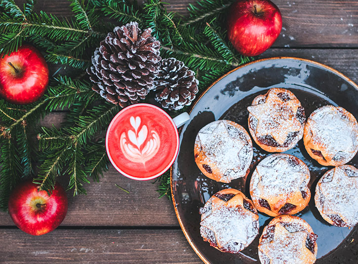 Festive cookies, a red latte, and red apples on a wooden table with pine boughs and pinecones