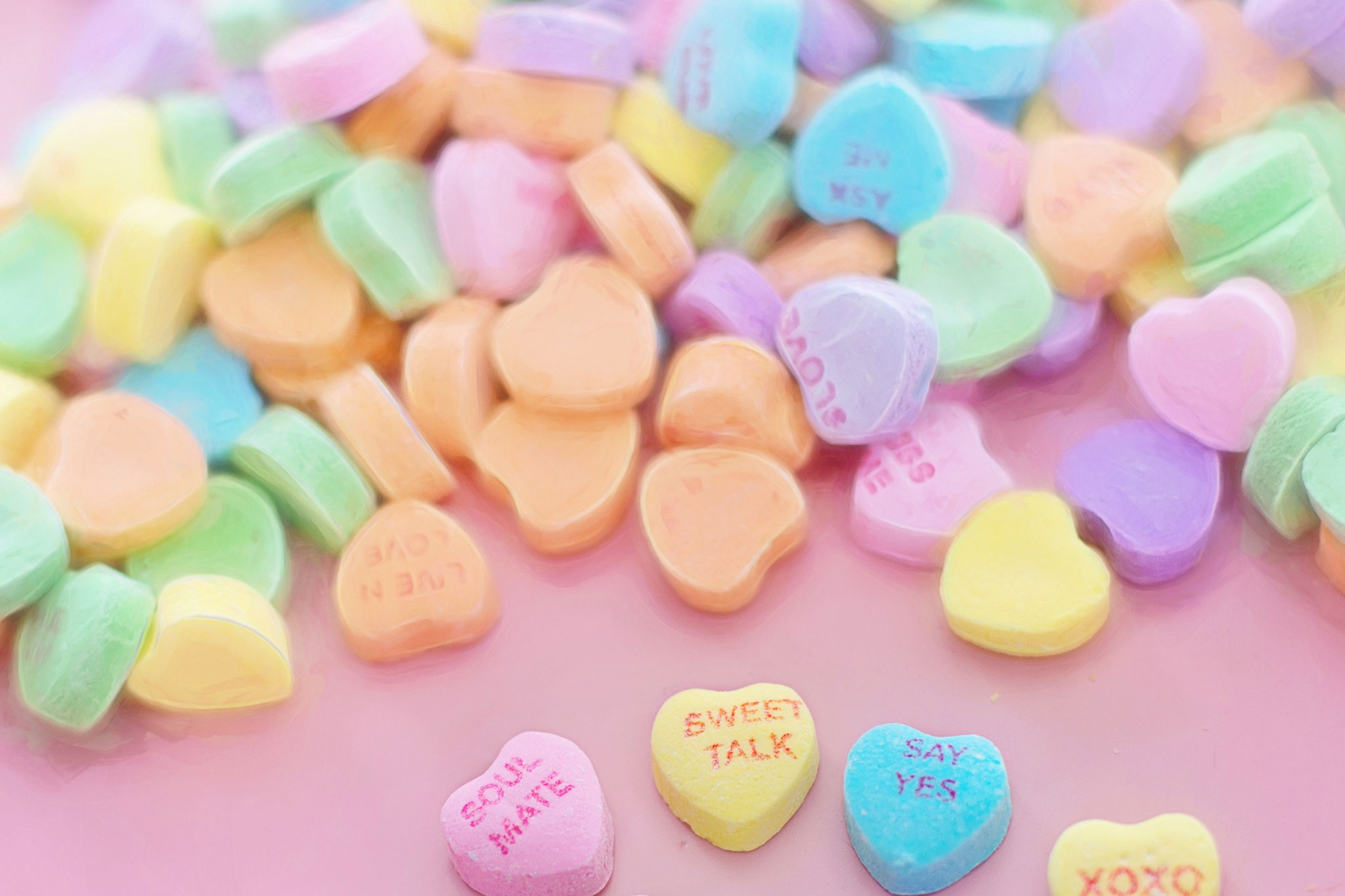 Multicolored Valentine's Day candy hearts with messages written on them scattered on a pink background