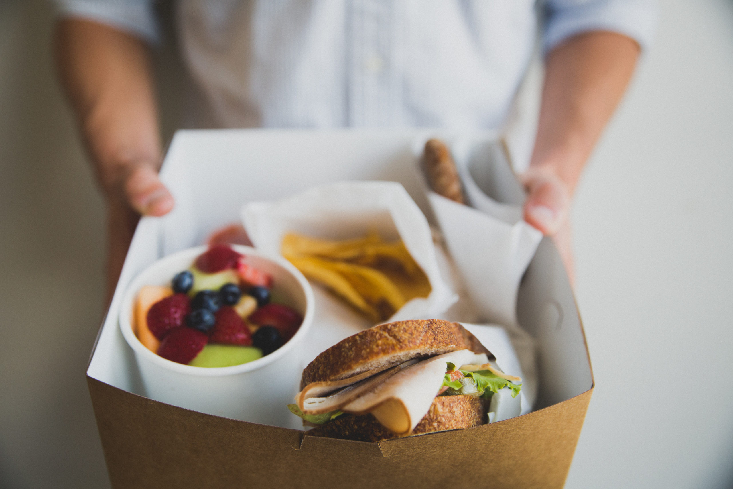 Someone holds out a boxed lunch with a sandwich, fresh fruit cup, and snack inside