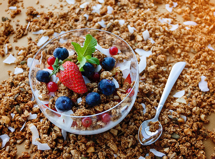 A bowl of granola, yogurt, and fruit sit on top of granola spread out on a board