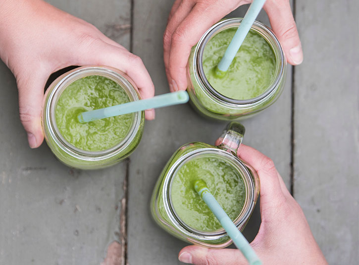Three hands hold cups full of green smoothies with straws in them