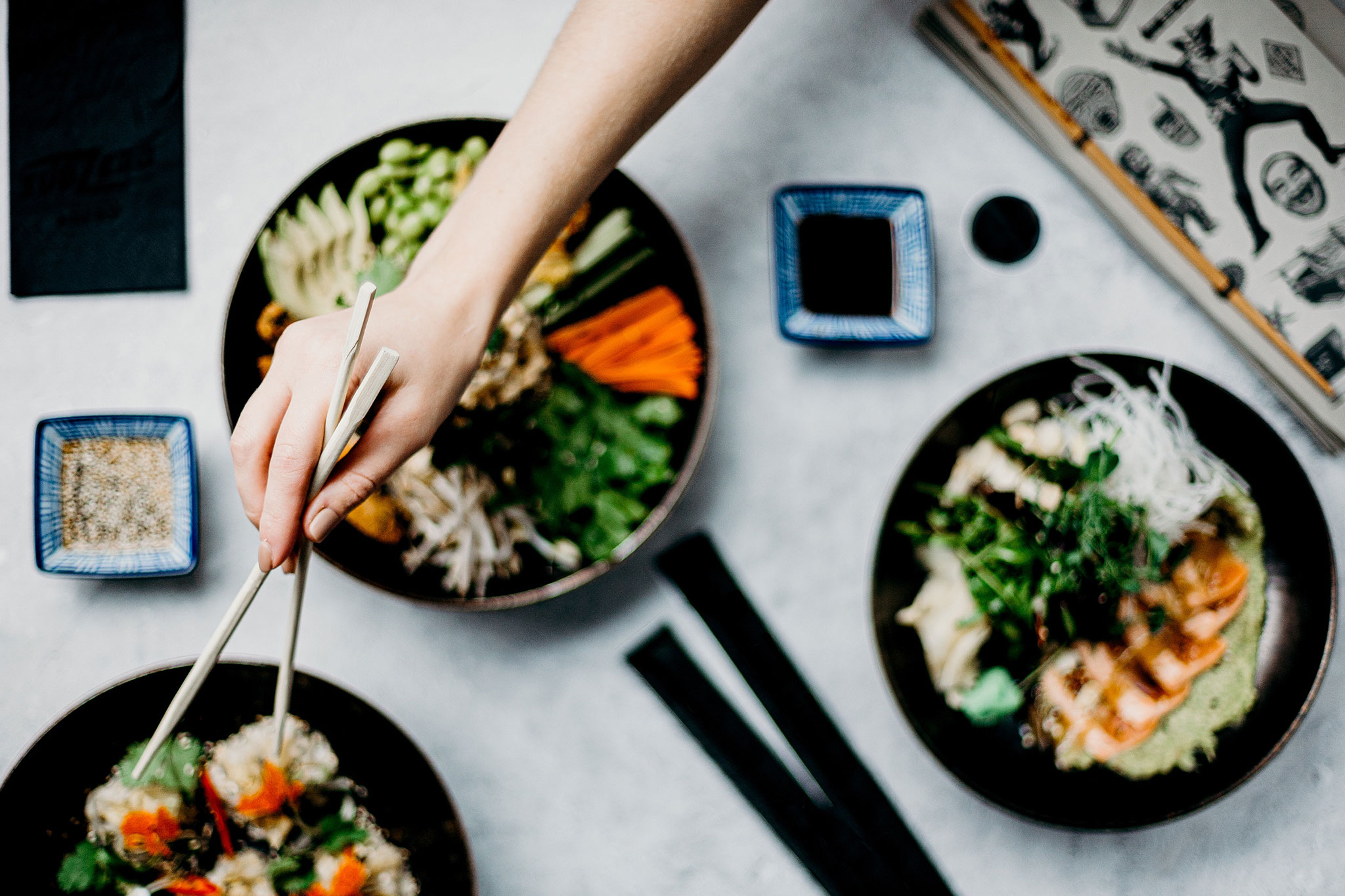 Hands holding chopsticks reach over sushi platters and soy sauce bowls to pick up a roll