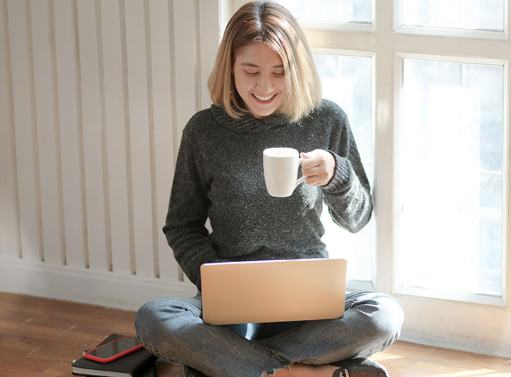 A woman sits crosslegged on the floor with her laptop in her lap and a coffee in her hand