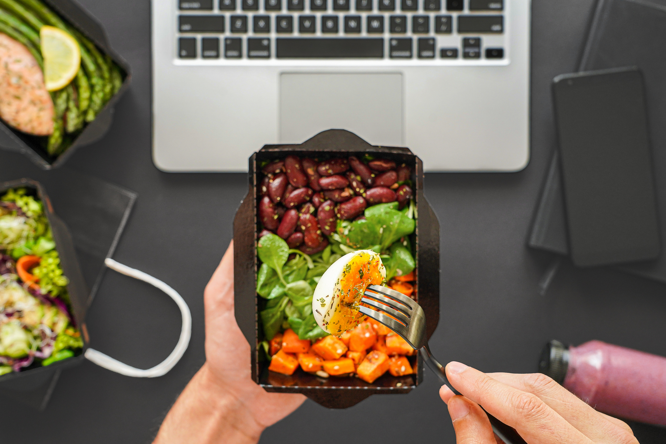 Hands hold a fork with an egg on it from a healthy takeout lunch over a laptop