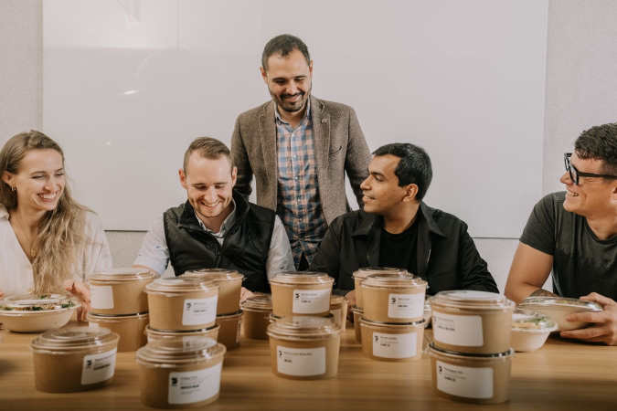 5 professionals sitting at a table and picking out their individually packaged Foodee order from a table filled with other Foodee orders.