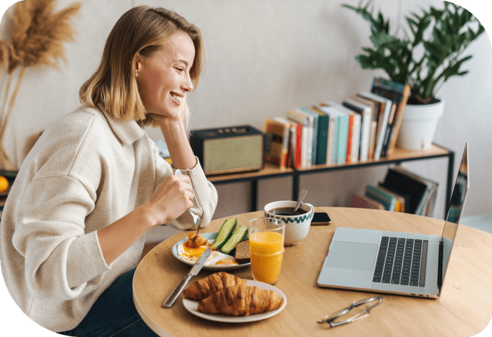 A woman sitting at a desk with a plate of croissants, sliced avocado, eggs, coffee and orange juice.