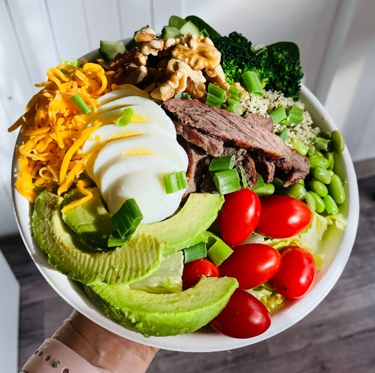 A person holding up a Rocket Bowl containing Quinoa, hard-boiled egg, avocado, chickpeas, shredded carrots, broccoli, cabbage, tomatoes, green onions & chia seeds.