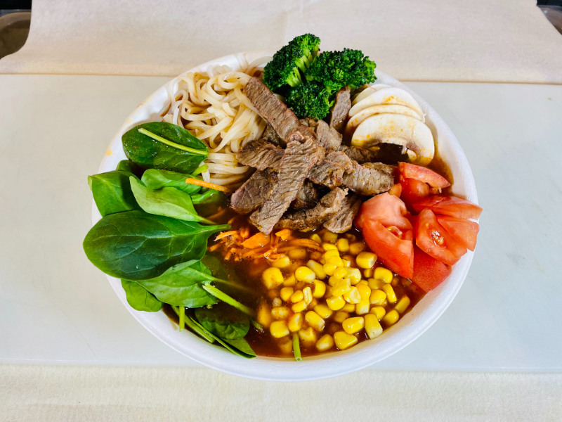 A bowl of Spicy Beef Noodle Soup with steak, spinach, carrots, corn, tomatoes, broccoli, mushrooms, green onion, rice noodles, beef Tom Yum broth.