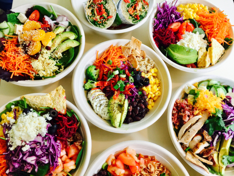 Seven bowls of salad on a table each with vibrantly coloured ingredients like shaved carrots and cabbage and sliced avocados and peppers.
