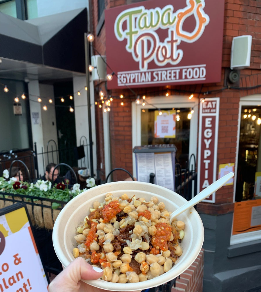 The front entrance of the Fava Pot restaurant with an individual holding up a bowl of their ordered food.