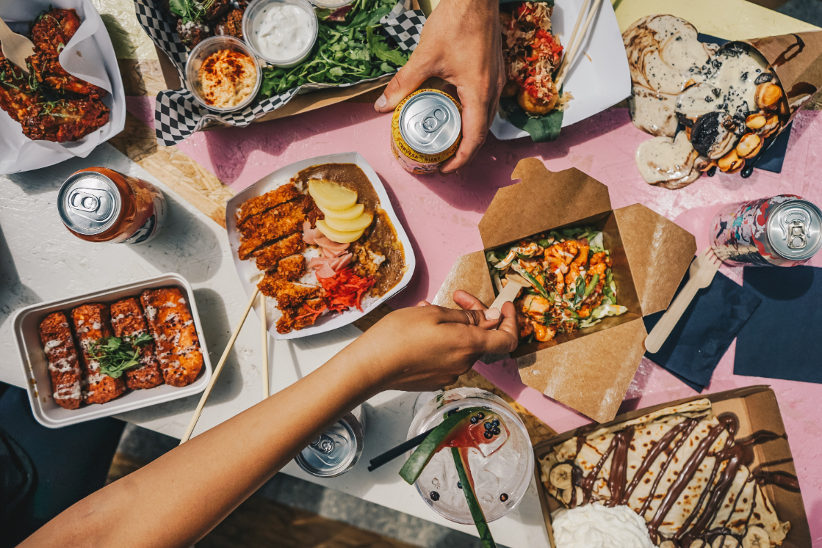 Hands reaching for one of many dishes in open to-go containers on a table scattered with sodas and napkins