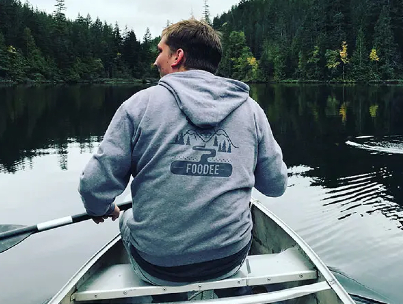 A Foodee employee wearing a grey hoodie with the Foodee logo on the back, sitting in a canoe on a Lake with green trees in the background.