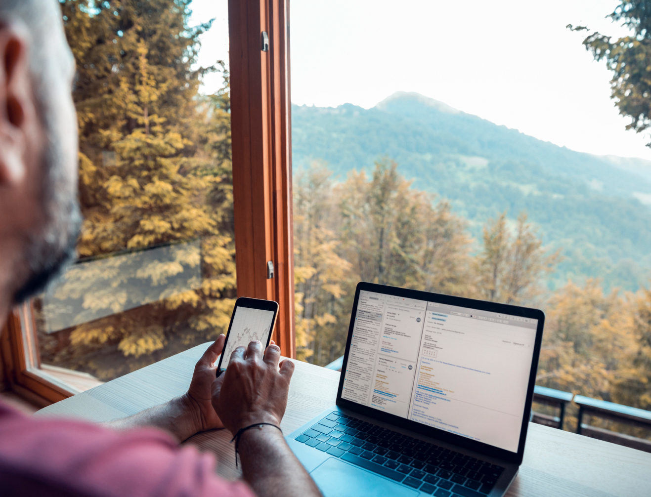 A individual working on their computer and phone in front of a large window overlooking the mountain and treetops.