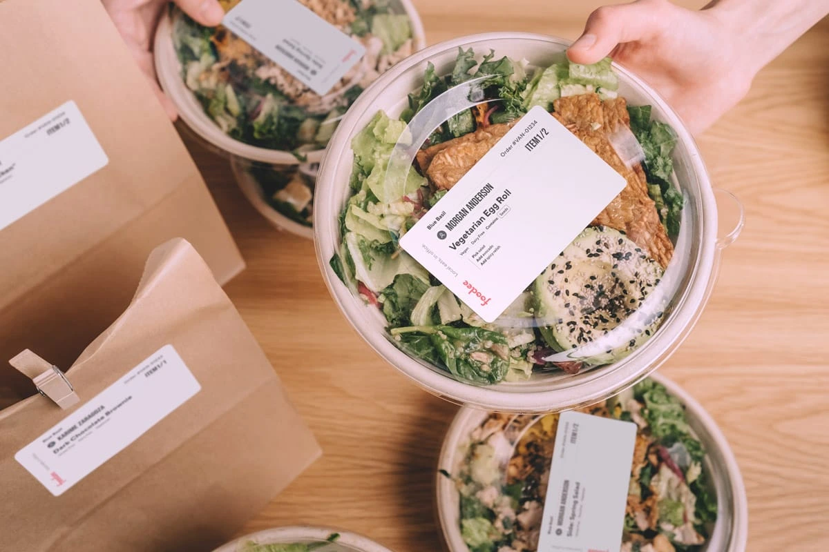 A hand grabbing one of three labeled to-go containers with salad inside next to two labeled brown paper bags.