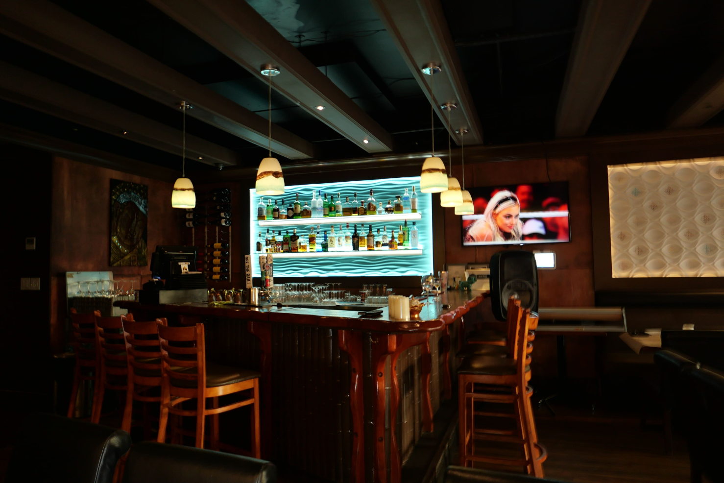Bombay's bar with a backlit shelf containing alcohol surrounded by a square shaped countertop with wooden barstools.