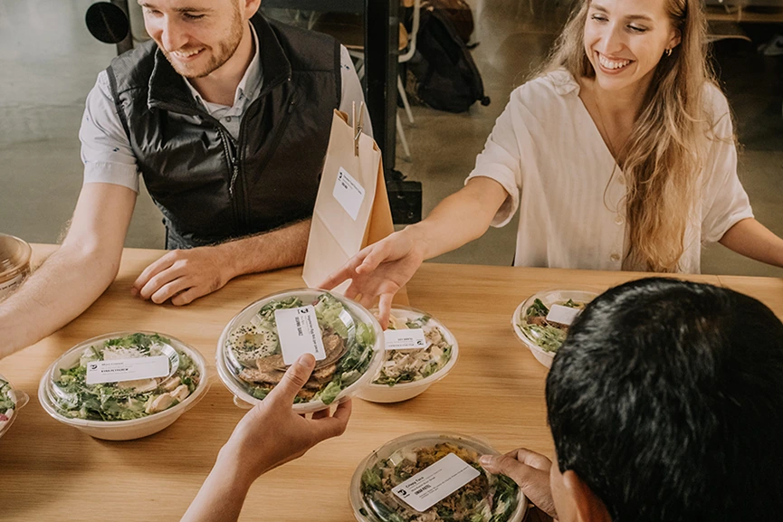 A group of coworkers sitting around a square table, distributing packaged foods to each other.