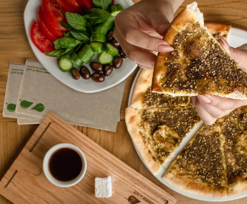 An individual holding a slide of the Zaatar flatbread, with a side plate of vegetables and a drink.