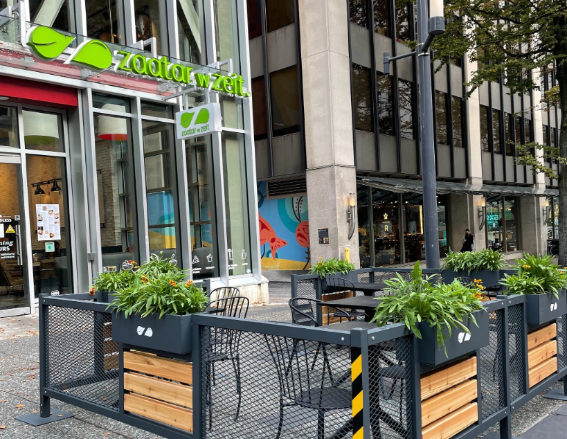 An enclosed patio at the front of the Zaatar W Zeit restaurant with black tables and chairs and decorative plants.