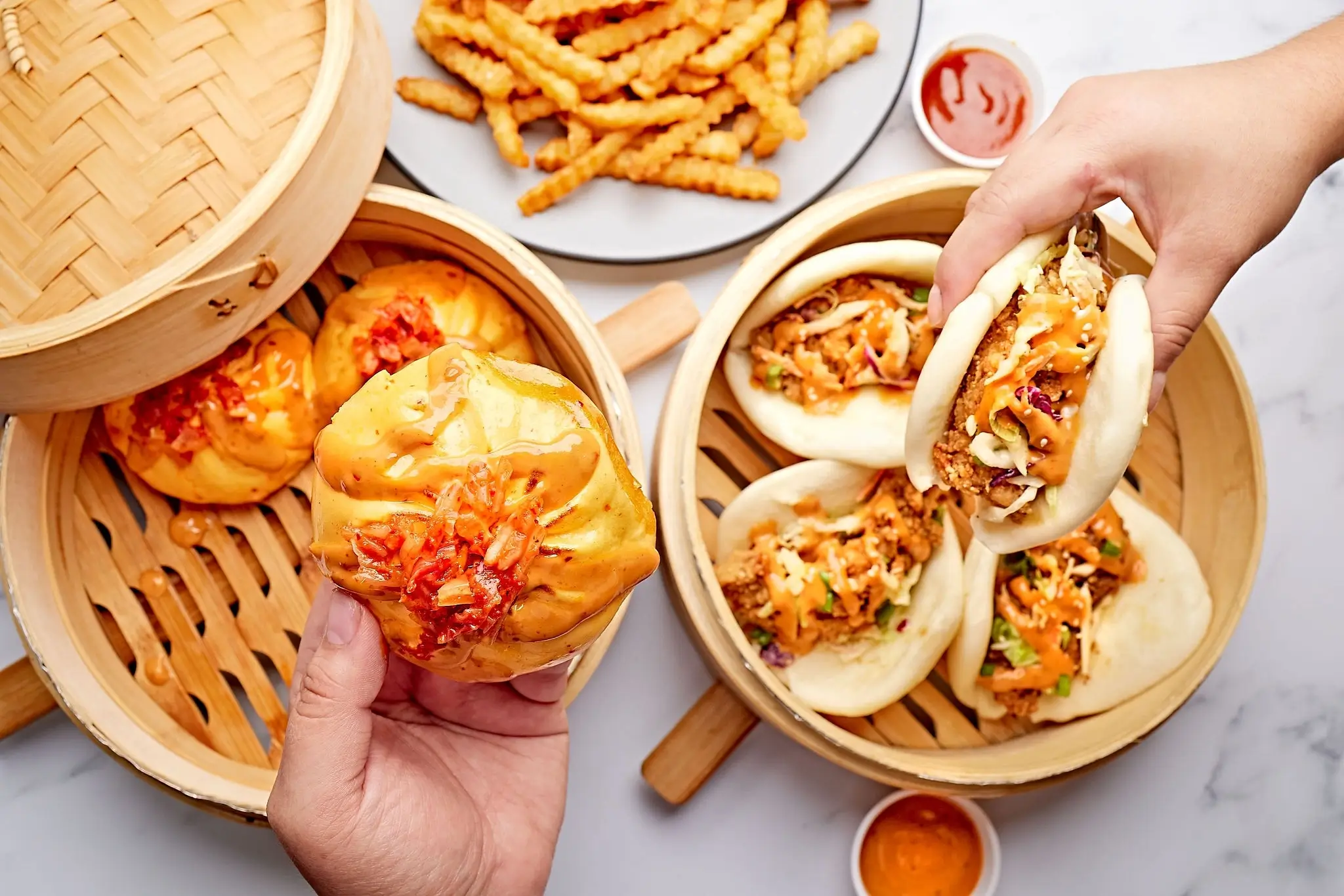 Hands reaching for a bao bun and dumpling from steamer baskets