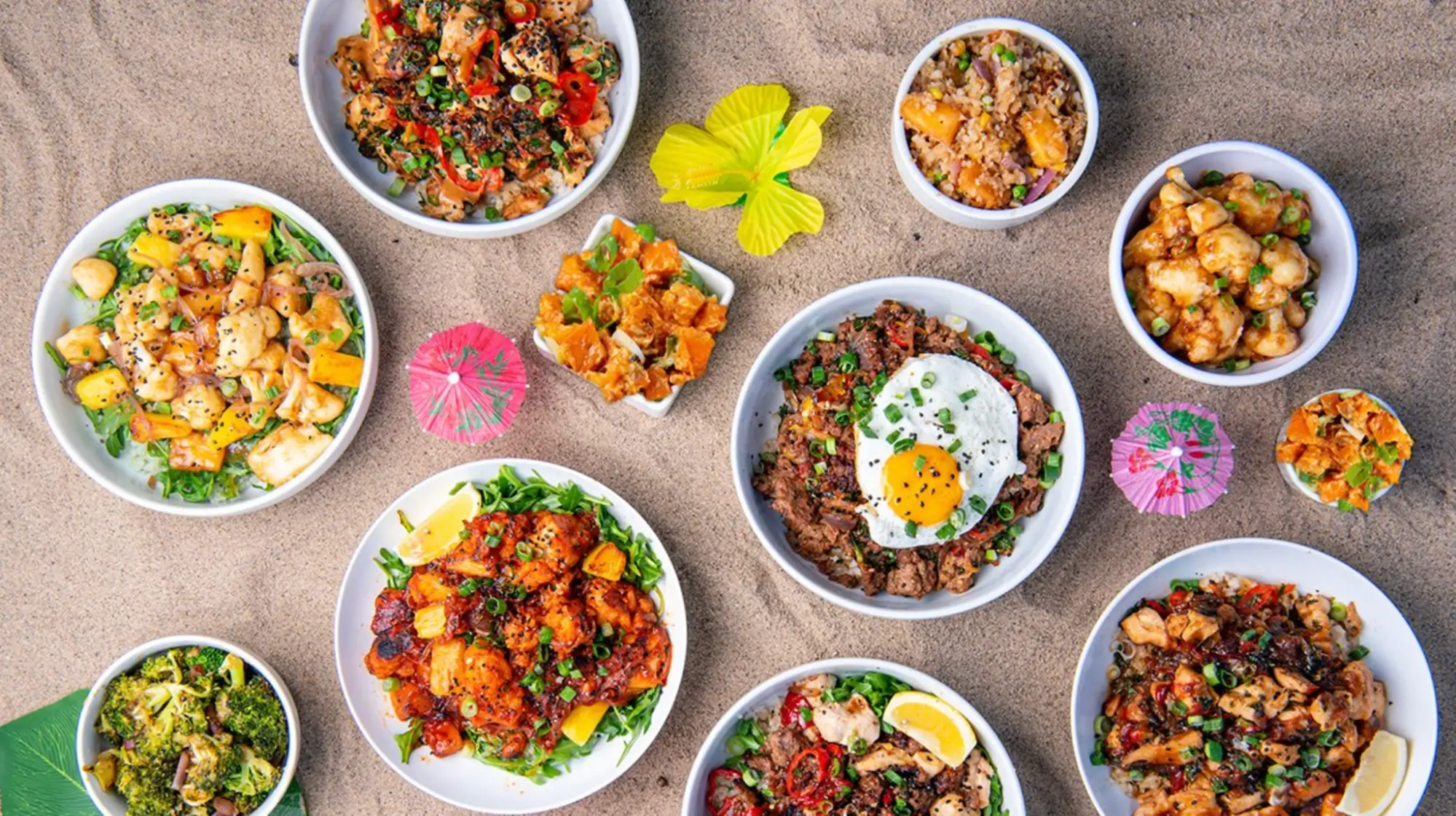 An assortment of bowls of Hawaiian-looking meals on a sandy background with a tropical flower and drink umbrellas