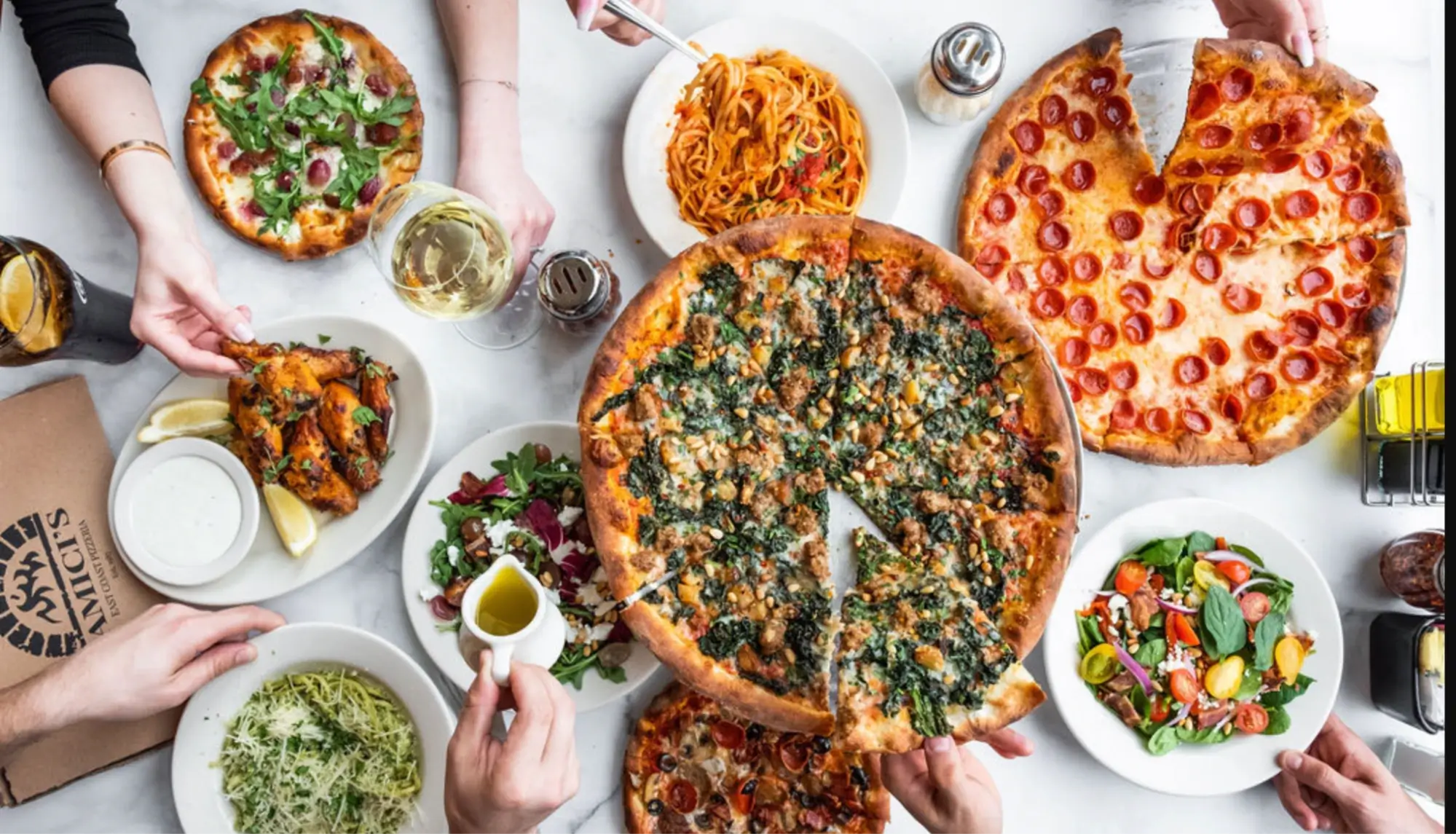 Hands reaching for slices of different pizzas, chicken wings, and salads on a table