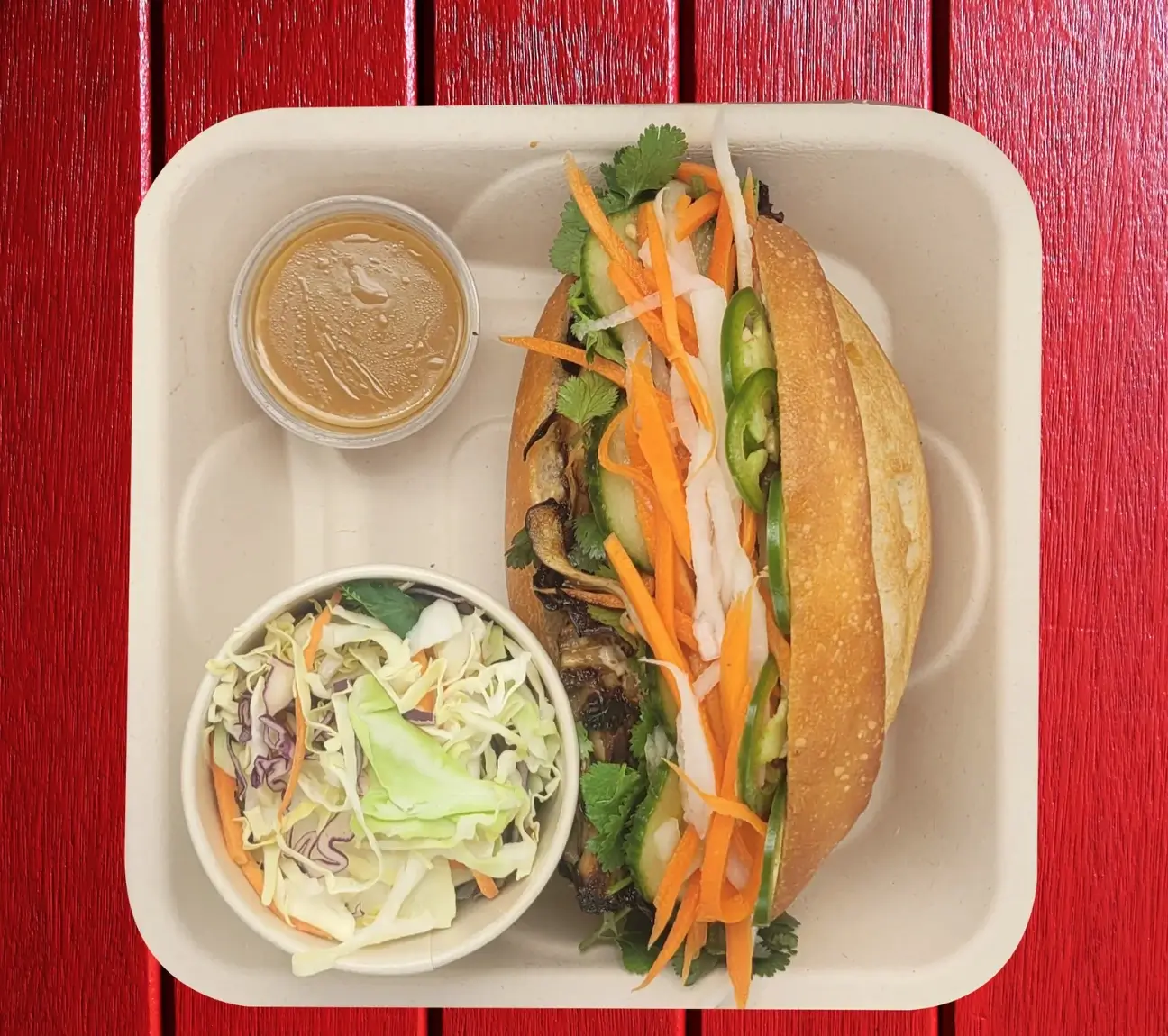 A Vietnamese-sub sandwich full of vegetables with a side coleslaw and sauce in a to-go container on a red background