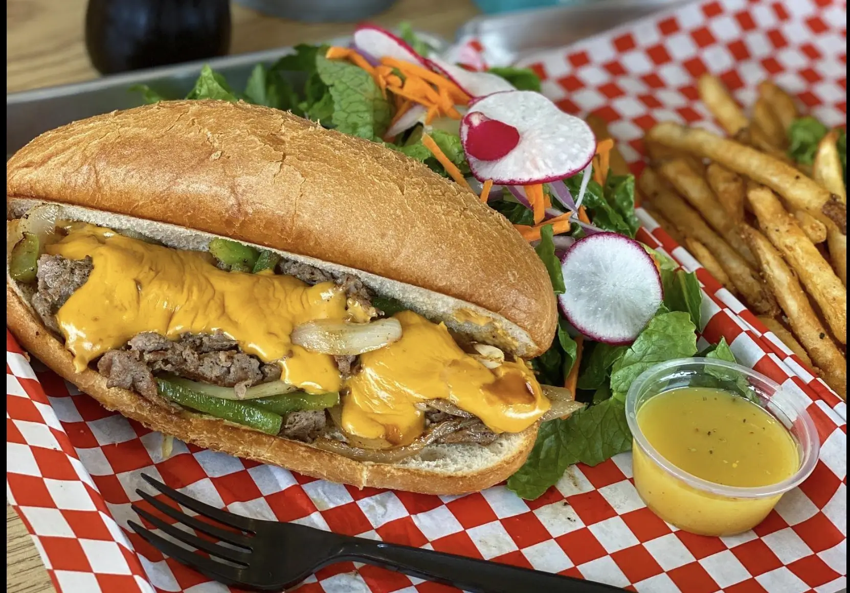 A sub sandwich with melted cheese and meat, a side salad, dressing, and fries on red and white checkered paper