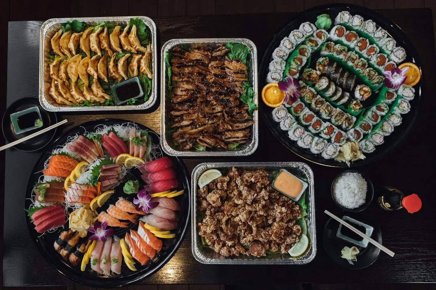 Large platters of assorted sushi, potstickers, and grilled meats with soy sauce and chopsticks on the side