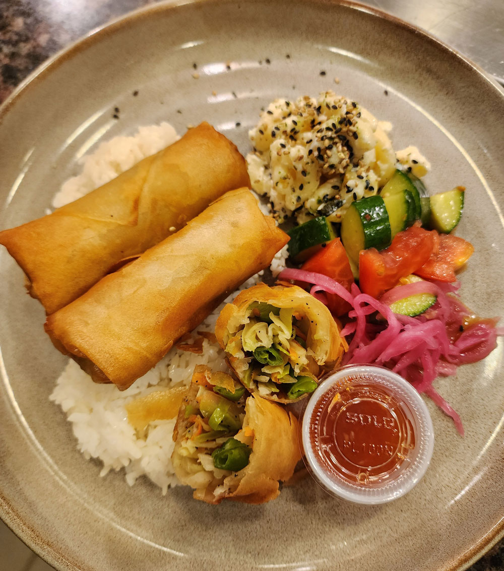 A plate of white rice topped with four spring rolls, sweet and sour sauce and chopped cucumbers.