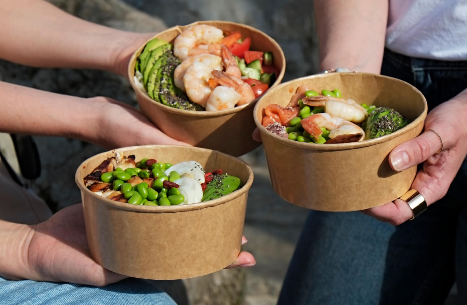 Three people holding up food bowls with beans, avocado and shrimp.